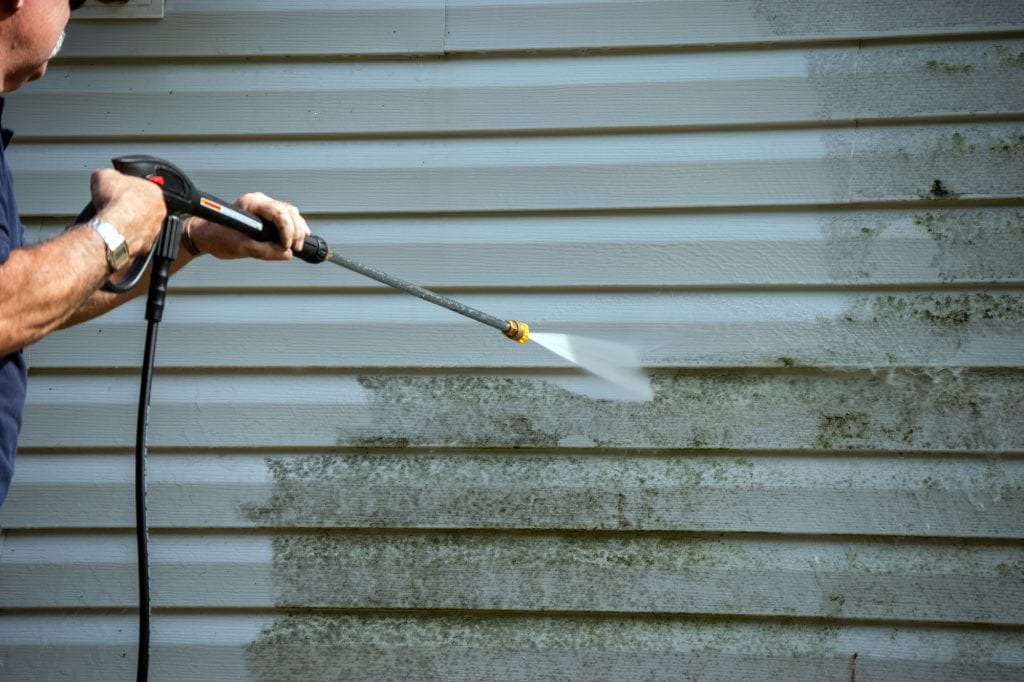 Man uses a power washer to clean mold and grime off the siding of a house.